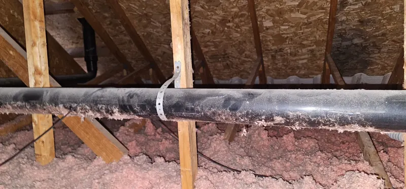 Grenier avant les travaux : ancien isolant en vrac rose, inégal, sous un gros tuyau de plomberie noir, entre les fermes de toit