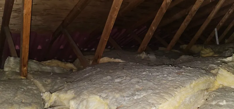Grenier avant les travaux : anciennes nattes jaunies et isolant en vrac gris, tassés entre les fermes de toit, déflecteurs de ventilation au fond
