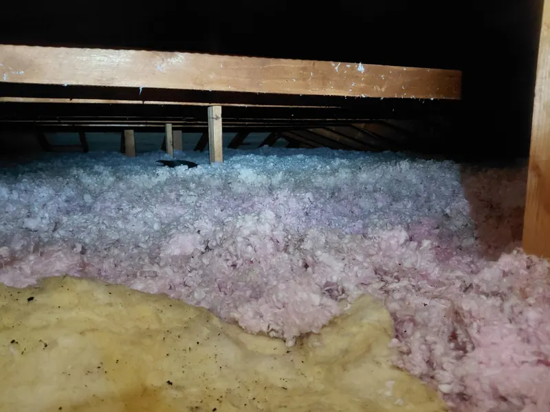 Grenier avant les travaux : ancienne laine soufflée rose posée sur une vieille natte jaune salie, vue au ras de l'isolant sous la charpente