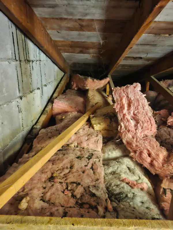 Grenier avant les travaux : vieilles nattes de laine rose et jaune, affaissées et salies, entre les solives, le long d'un mur de blocs de béton
