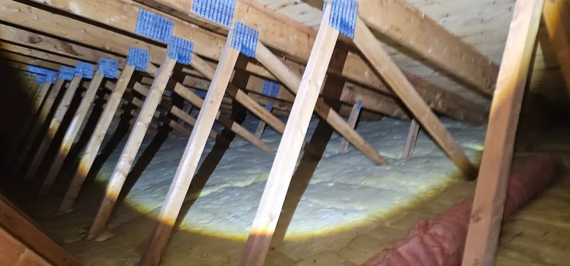 Grenier avant les travaux : ancien isolant jaunâtre sous une longue rangée de fermes de toit, éclairé à la lampe de poche